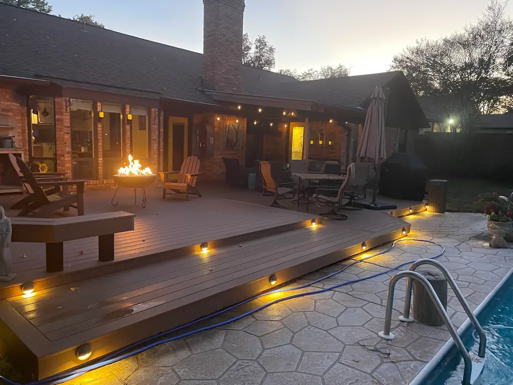 Composite deck at dusk with lit riser lights, a fire bowl and a pool beyond